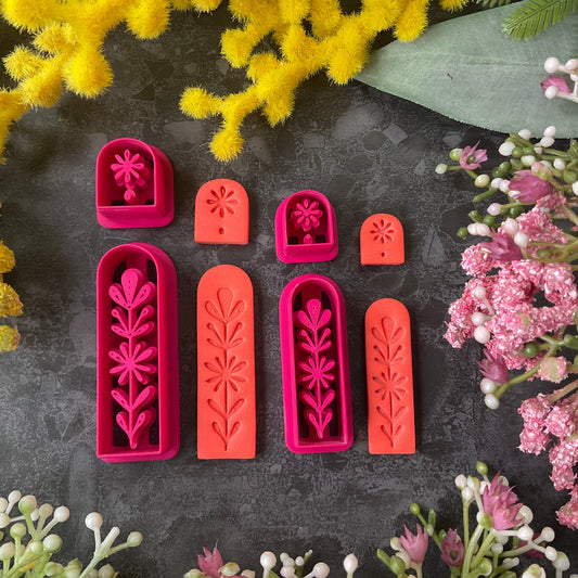 A set of 3d printed flower-shaped polymer clay cutters in different sizes arranged on a dark textured surface with a black ruler below and yellow and pink flowers around the edges. The cutters are hot pink and orange, Winter Meadow Polymer Clay Cutter Set.