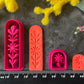 A set of 3d printed flower-shaped polymer clay cutters in different sizes arranged on a dark textured surface with a black ruler below and yellow and pink flowers around the edges. The cutters are hot pink and orange, Winter Meadow Polymer Clay Cutter Set.