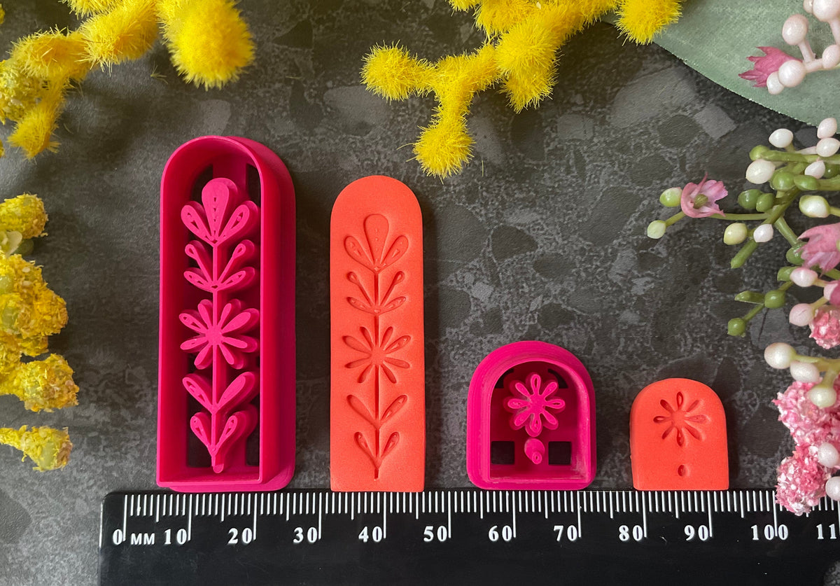 A set of 3d printed flower-shaped polymer clay cutters in different sizes arranged on a dark textured surface with a black ruler below and yellow and pink flowers around the edges. The cutters are hot pink and orange, Winter Meadow Polymer Clay Cutter Set.