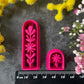 A set of 3d printed flower-shaped polymer clay cutters in different sizes arranged on a dark textured surface with a black ruler below and yellow and pink flowers around the edges. The cutters are hot pink and orange, Winter Meadow Polymer Clay Cutter Set.