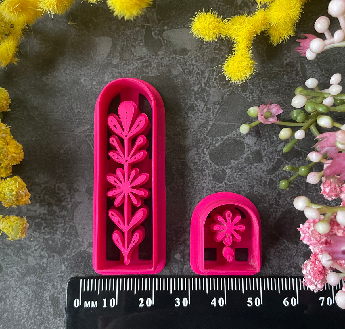 A set of 3d printed flower-shaped polymer clay cutters in different sizes arranged on a dark textured surface with a black ruler below and yellow and pink flowers around the edges. The cutters are hot pink and orange, Winter Meadow Polymer Clay Cutter Set.