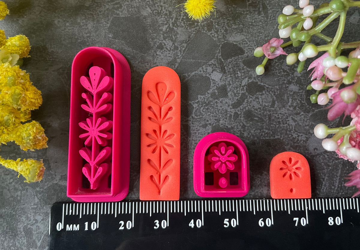 A set of 3d printed flower-shaped polymer clay cutters in different sizes arranged on a dark textured surface with a black ruler below and yellow and pink flowers around the edges. The cutters are hot pink and orange, Winter Meadow Polymer Clay Cutter Set.