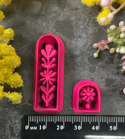 A set of 3d printed flower-shaped polymer clay cutters in different sizes arranged on a dark textured surface with a black ruler below and yellow and pink flowers around the edges. The cutters are hot pink and orange, Winter Meadow Polymer Clay Cutter Set.