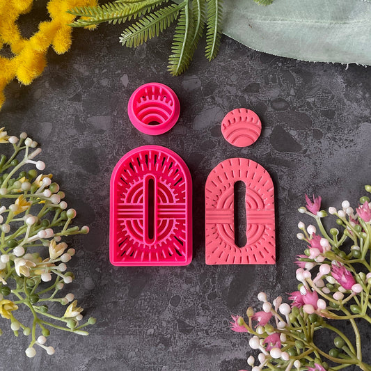 Bright pink 3d printed polymer clay cutters shaped like stylized  arch and circular figures, displayed on a dark textured surface surrounded by decorative flowers and greenery. Woven Earth Polymer Clay Cutter Set