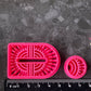 Bright pink 3d printed polymer clay cutters shaped like stylized  arch and circular figures, displayed on a dark textured surface surrounded by decorative flowers and greenery. Woven Earth Polymer Clay Cutter Set