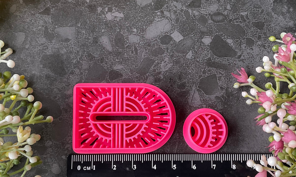 Bright pink 3d printed polymer clay cutters shaped like stylized  arch and circular figures, displayed on a dark textured surface surrounded by decorative flowers and greenery. Woven Earth Polymer Clay Cutter Set